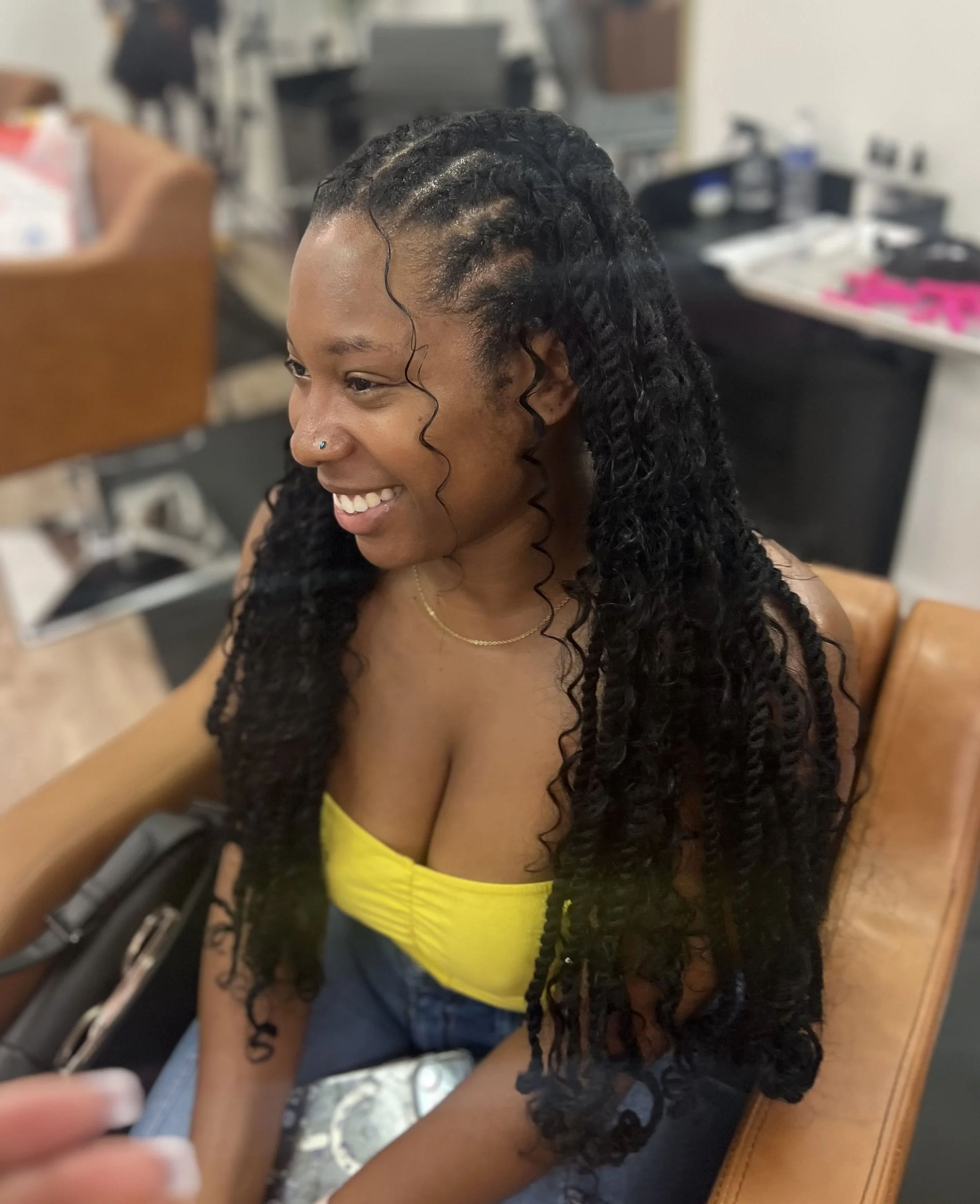Smiling woman with long curly hair, yellow top, sitting on a brown chair, in a room with various items in the background.