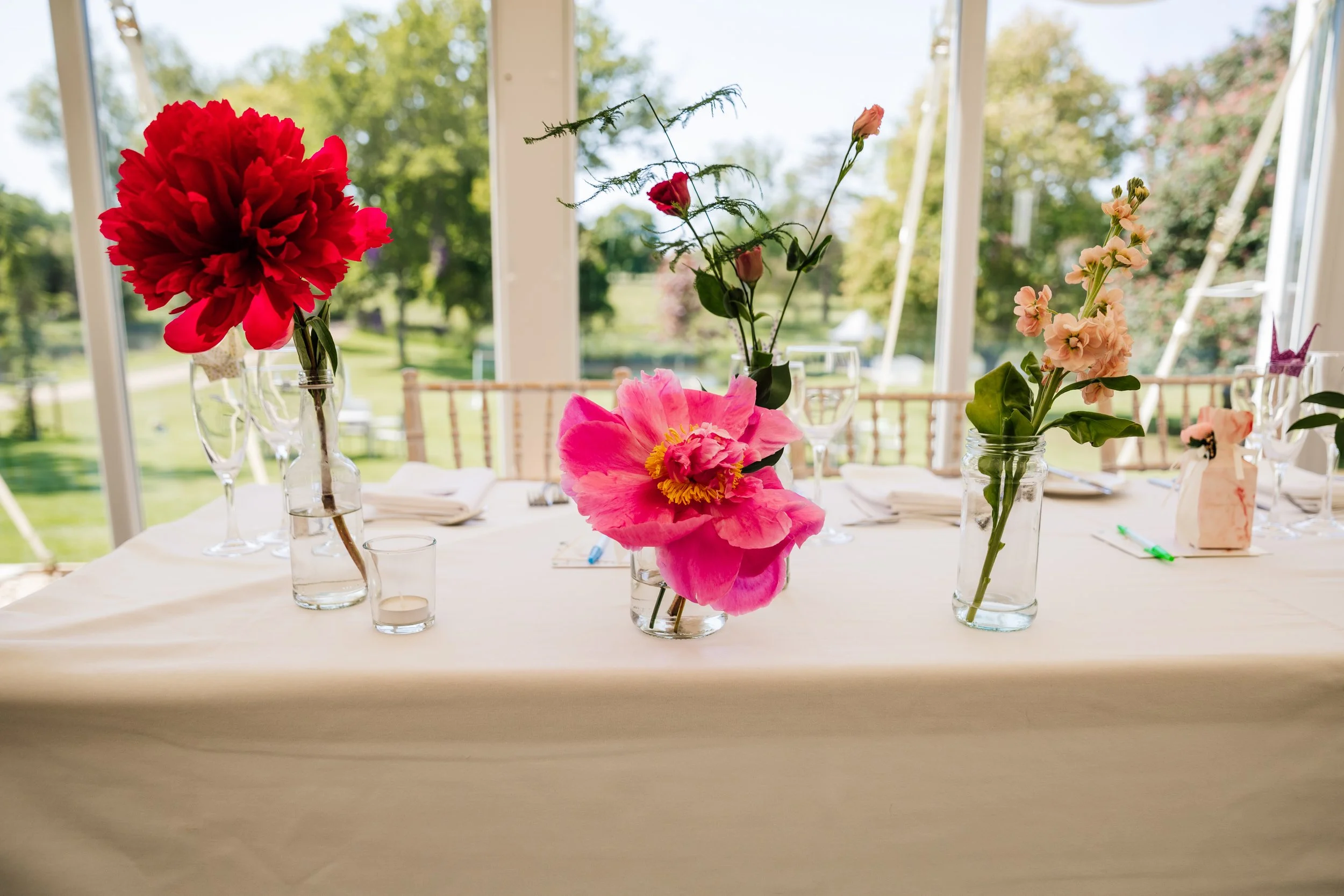 A dining table decorated with colorful flowers in glass jars, set in a bright sunlit room with large windows showing outdoor greenery.