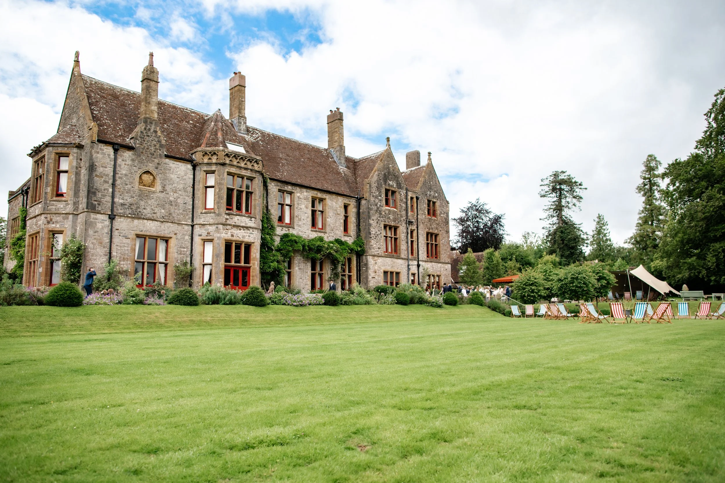 A historic stone mansion surrounded by a lush green lawn with garden chairs and trees, under a partly cloudy sky.