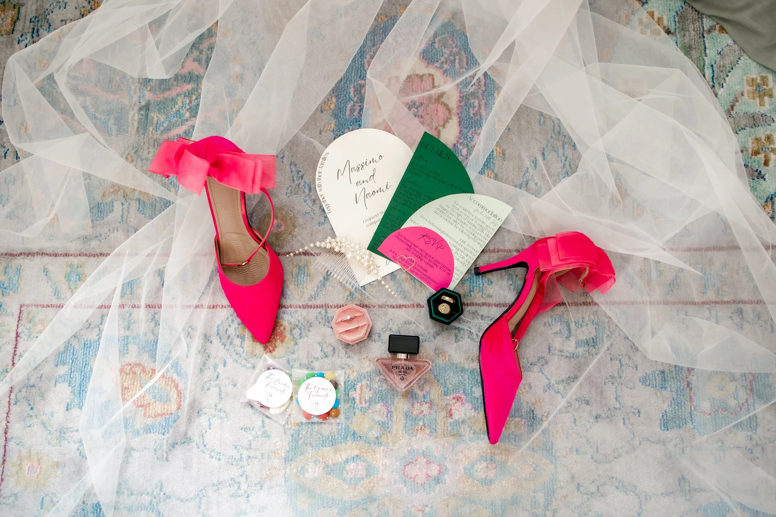 Pink high-heeled shoes with bows, wedding invitation, pearls, perfume, and candy on a colorful rug with a sheer white fabric overlay.