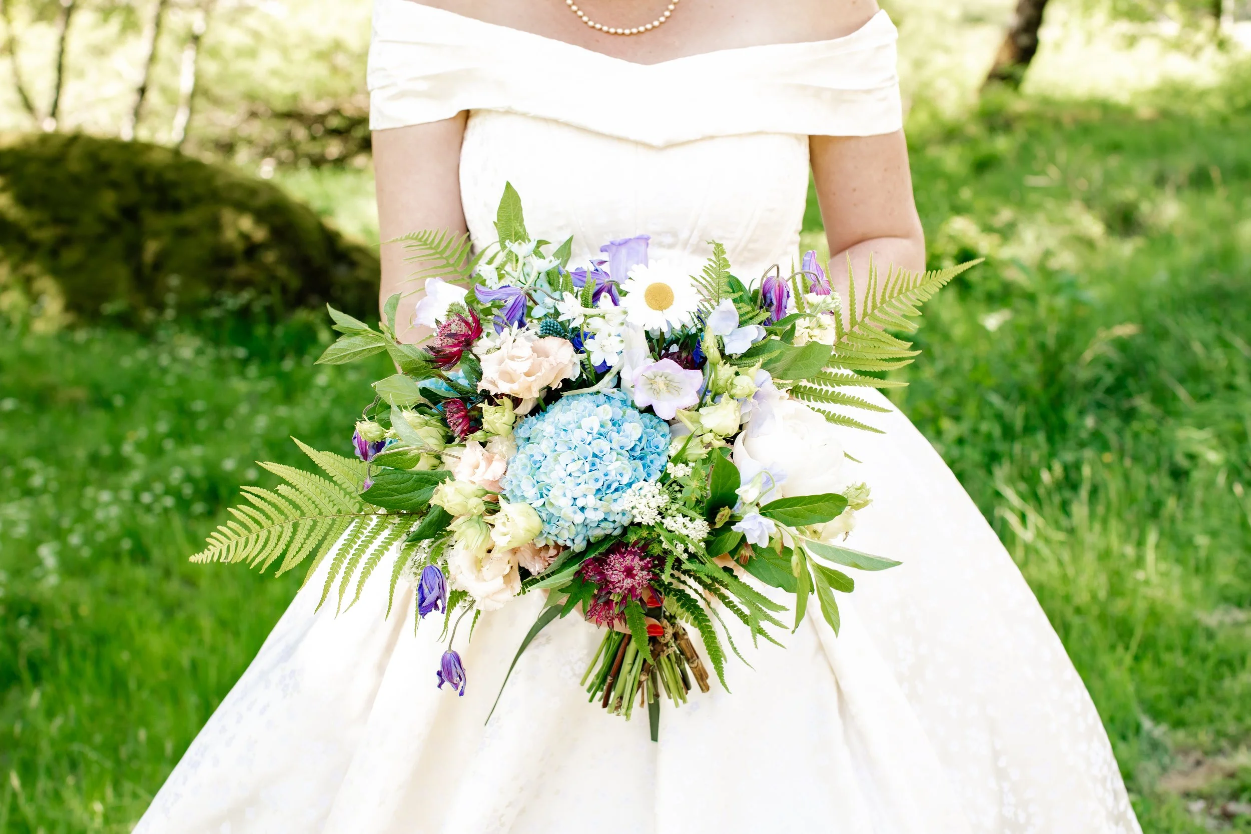 A bride in a white gown holding a colorful bouquet of flowers outdoors in a green garden.