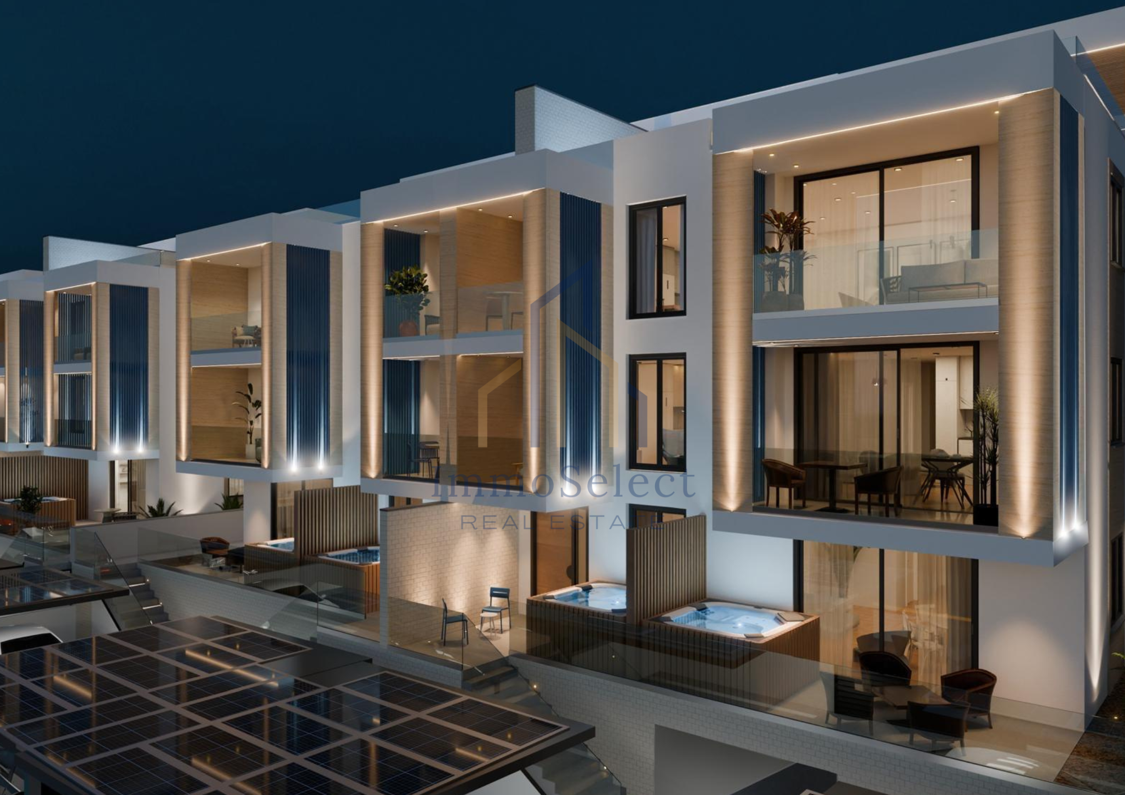 Night view of a modern multi-story residential building with illuminated balconies, large glass windows, and patio areas, featuring lighting accents and outdoor seating.