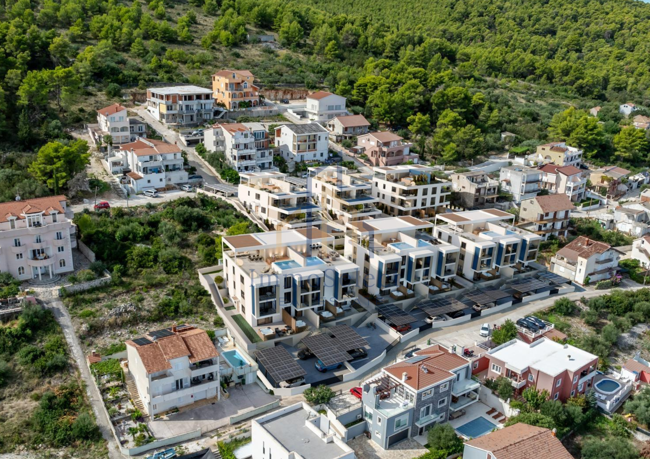 Aerial view of a residential neighborhood with modern white apartment buildings, parking areas with solar panels, neighboring houses, and lush green hills in the background.