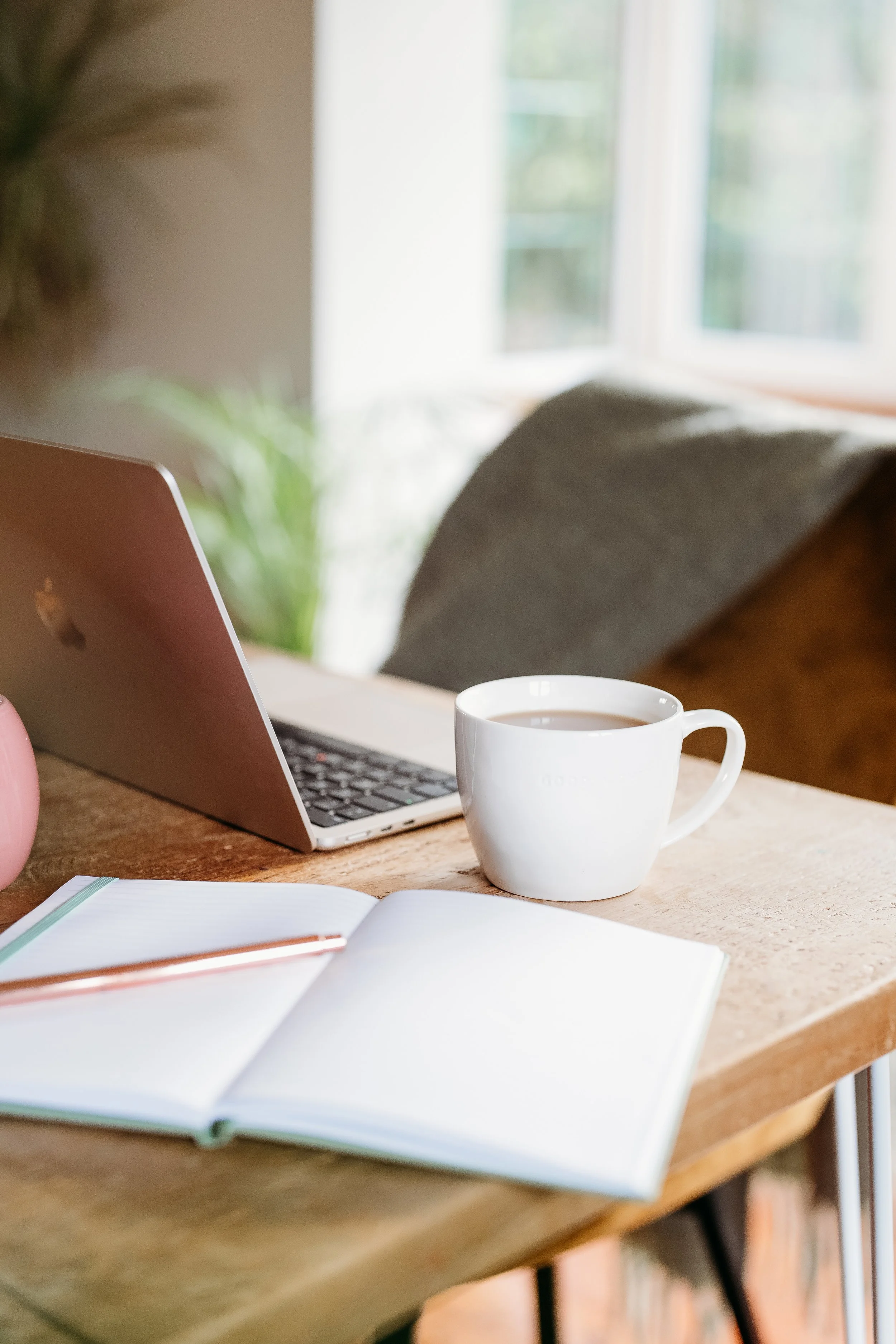 A wooden desk with a laptop, an open notebook with a pen, and a cup of coffee near a window.