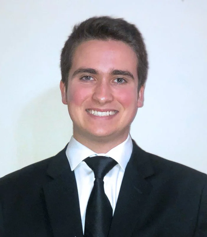 A young man in a black suit, white shirt, and black tie, smiling, with short brown hair, against a plain light-colored background.