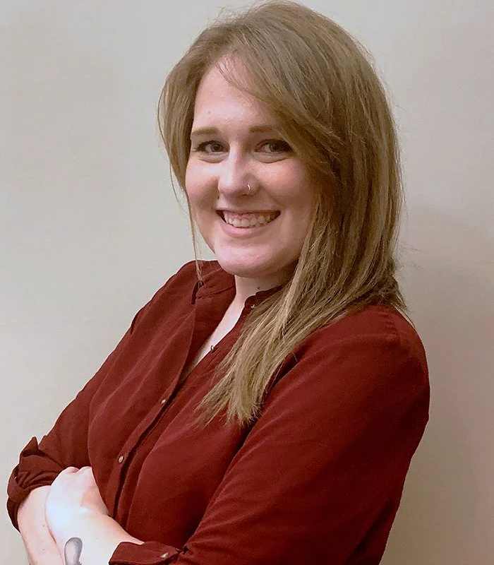 Laurel Kiernan headshot with long, light brown hair, wearing a nose ring and a red button-up shirt, standing against a plain beige wall.