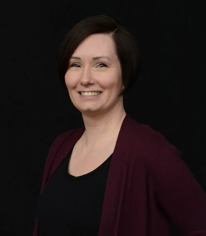 Tammi Anderson headshot with short dark hair, smiling, wearing a black top and a dark red cardigan, against a black background.
