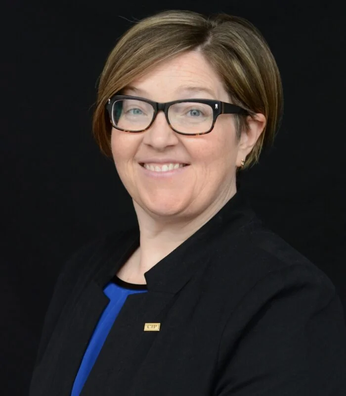 Amanda Farrell Walsh headshot with short light brown hair, glasses, and a black blazer smiling at the camera against a black background.