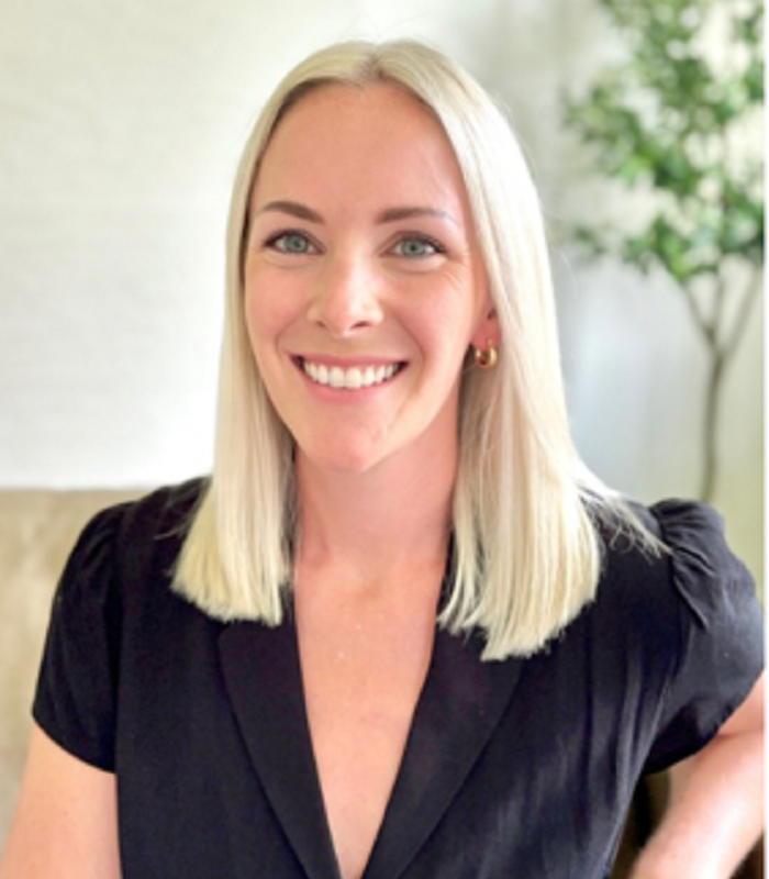 Laura Phillips headshot with straight blonde hair and blue eyes, wearing a black top with puffed sleeves, gold hoop earrings, and standing in front of a neutral background with a plant in the distance.