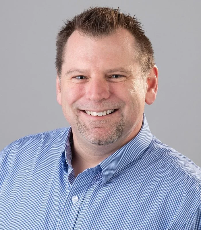 Manny Page headshot of a smiling middle-aged man with short brown hair, blue eyes, and a goatee, wearing a light blue checkered shirt against a plain gray background.