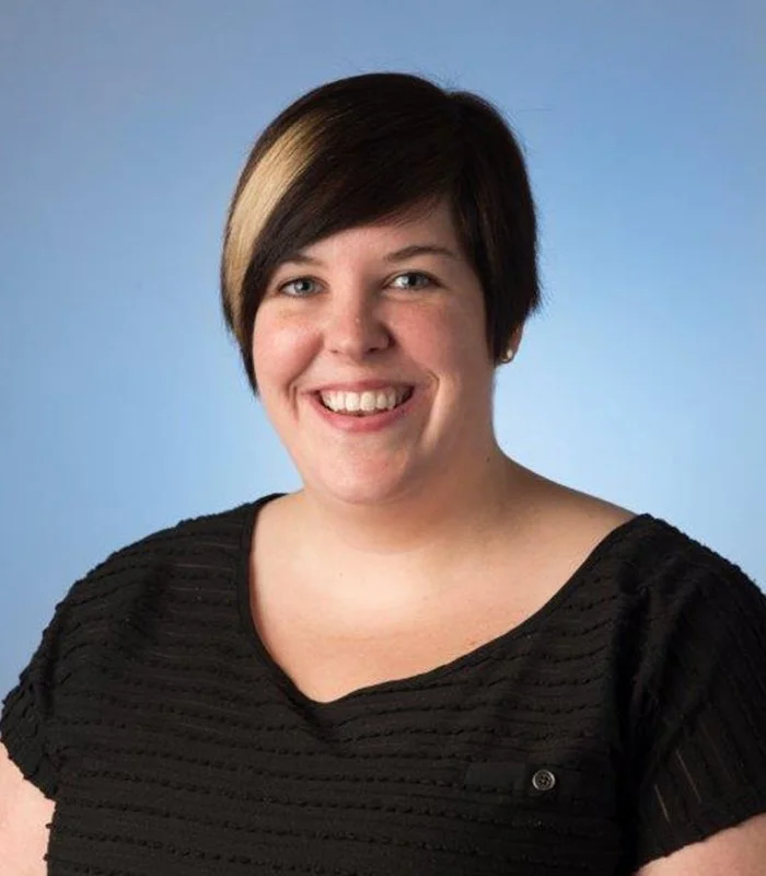Jessica Campbell headshot with short dark hair with a light streak, wearing a black textured top, against a blue background.