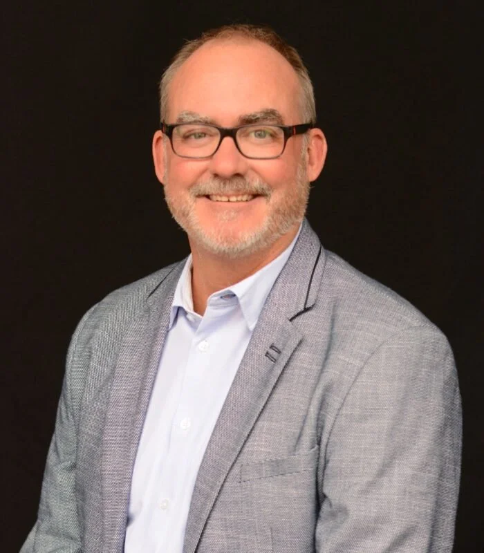 A middle-aged man with glasses, gray hair, and a beard, smiling, wearing a light gray blazer and white shirt against a black background.