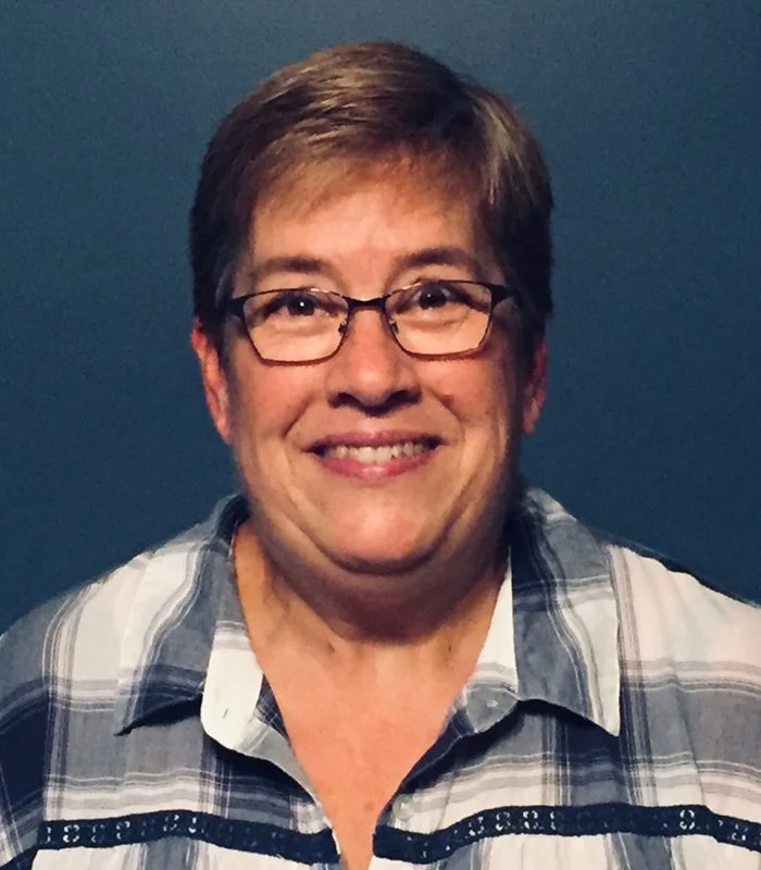 Gwen Liesemer headshot with short brown hair and glasses, smiling, wearing a blue and white plaid shirt against a dark background.