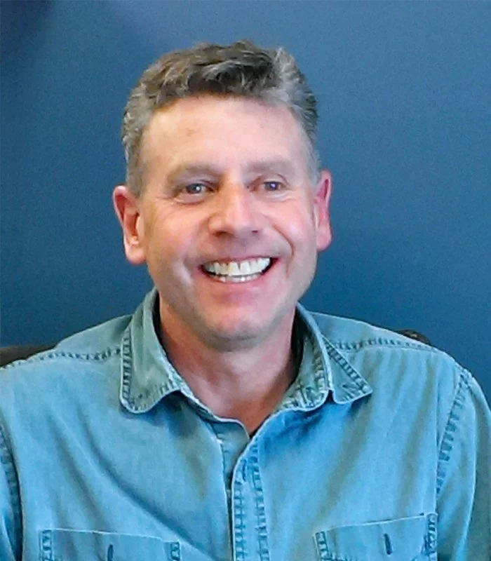 Jeff Chappel headshot with smiling man with short, curly gray hair wearing a light blue denim shirt against a dark background.