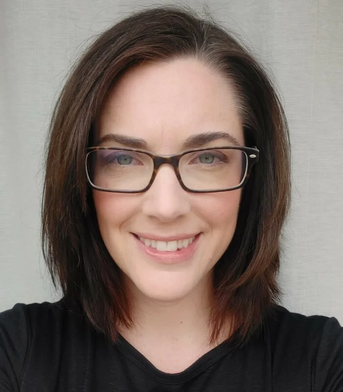 Tanya Coltman headshot with shoulder-length brown hair, glasses, and fair skin, smiling and wearing a black shirt, against a neutral background.