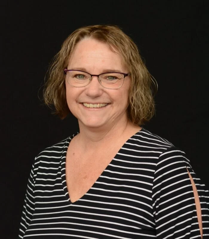 Carol Yoder head shot with short curly light brown hair, glasses, and a striped black and white shirt smiling against a black background.