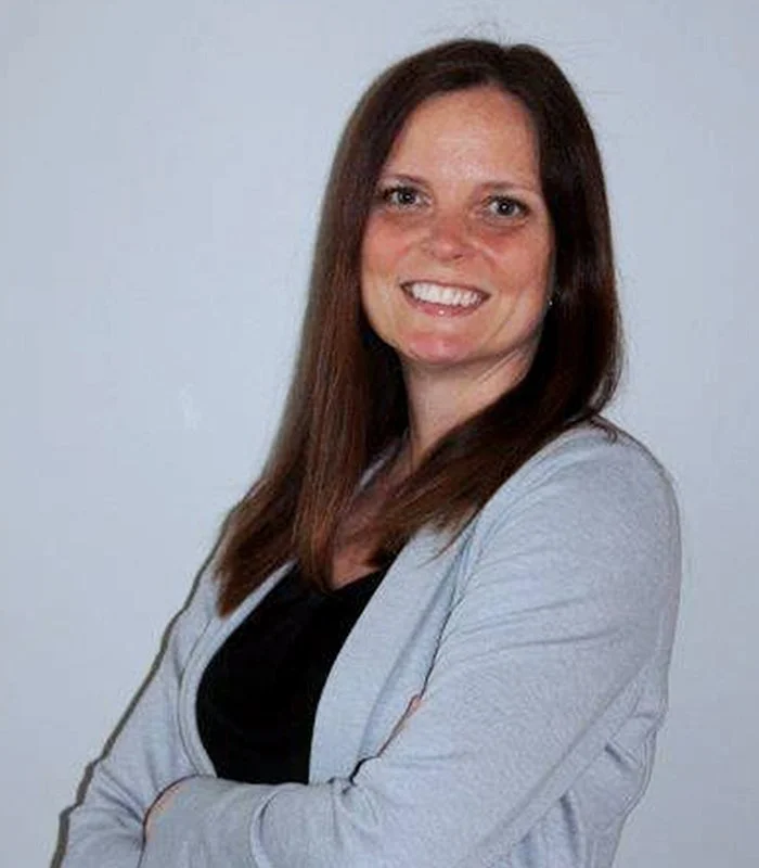 Laura Lambert headshot with long brown hair, smiling, wearing a black top and a light gray jacket, standing against a plain white background.