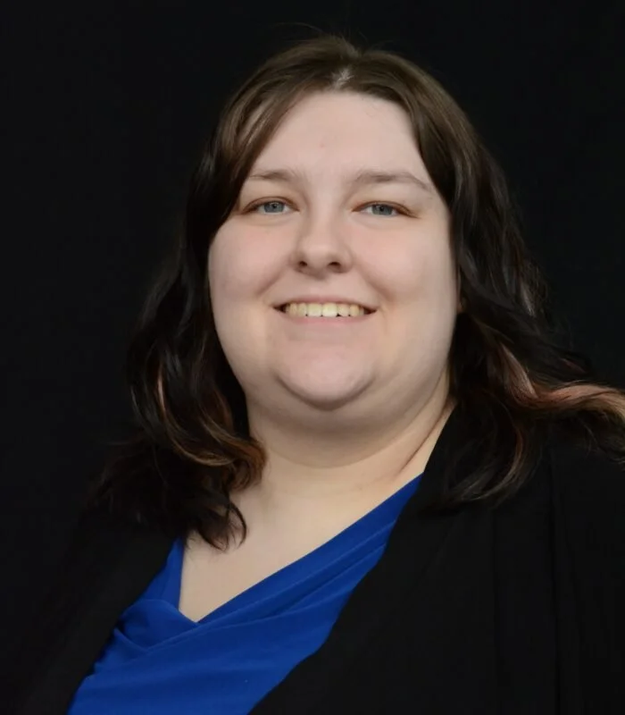 Alyssa Morris head shot with shoulder-length brown hair smiling, wearing a black blazer over a blue top, against a dark background.