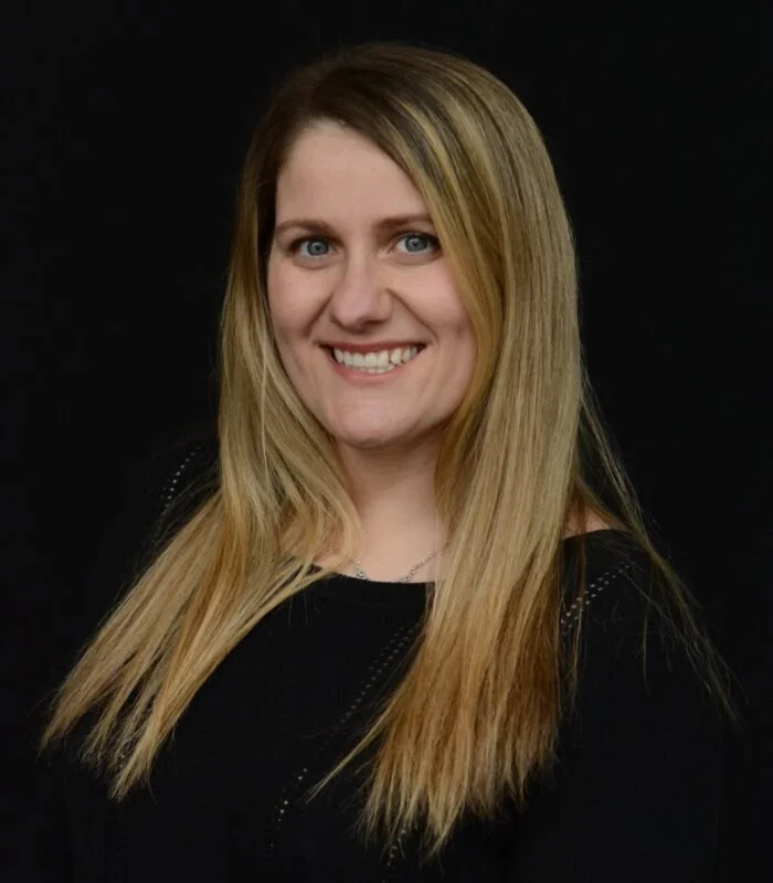 Portrait of a young woman with long blond hair and blue eyes, wearing a black top, smiling against a black background.