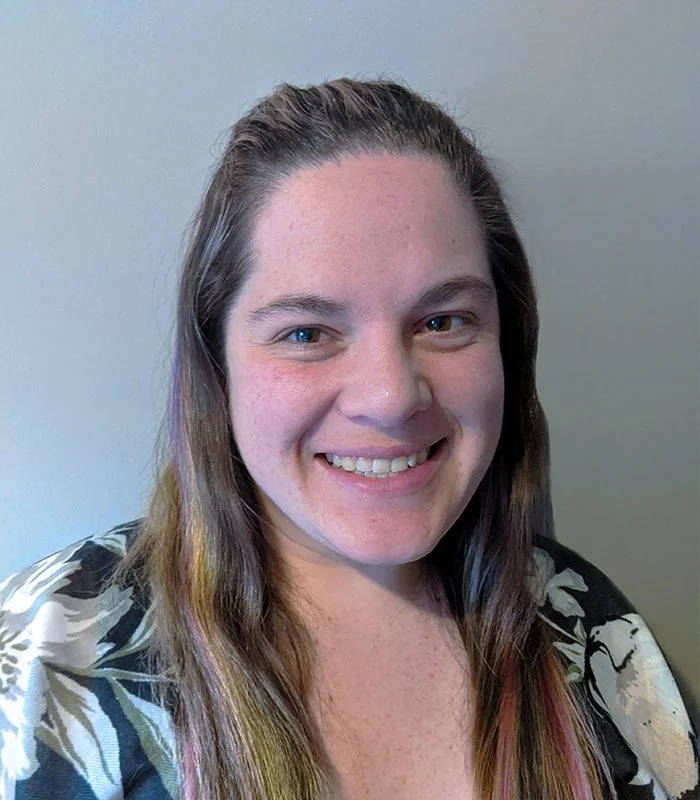 Close-up of a young woman with long brown hair and blue eyes, smiling at the camera, wearing a floral top, against a plain light-colored wall.