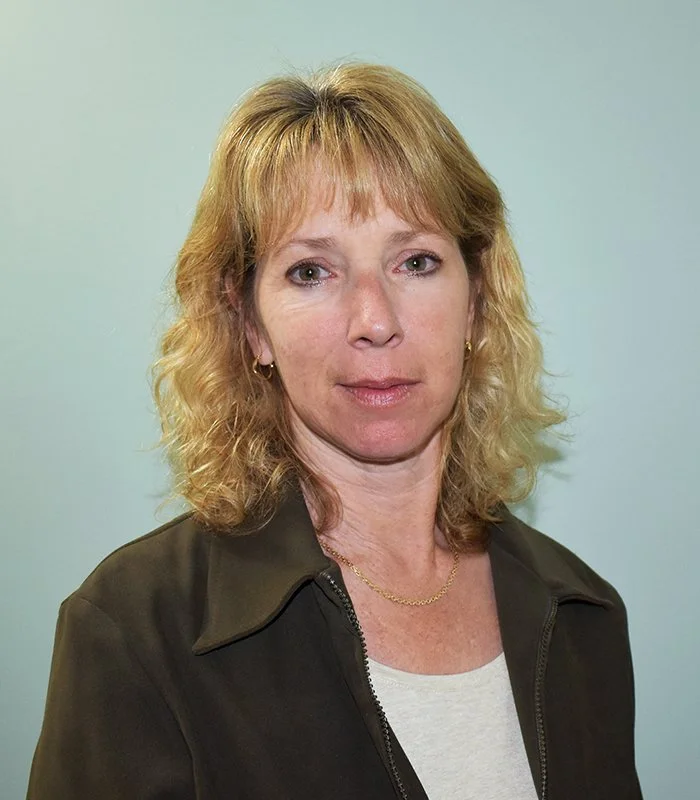 Lorraine Glover headshot with shoulder-length blonde curly hair, wearing a black jacket over a white top, standing against a light-colored background.
