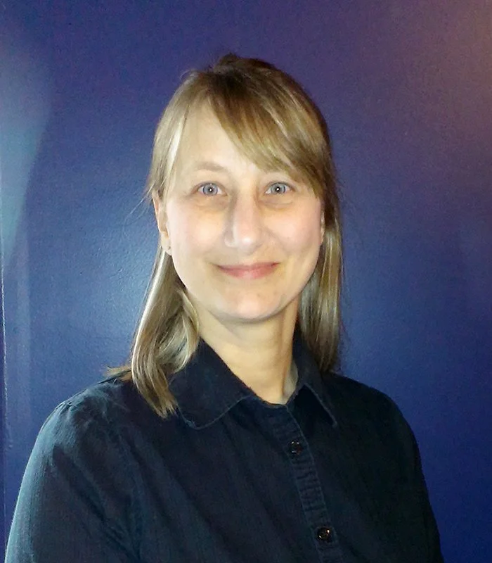 Debora Schaus headshot with shoulder-length blonde hair and blue eyes, wearing a dark button-up shirt, standing against a dark blue wall and smiling at the camera.