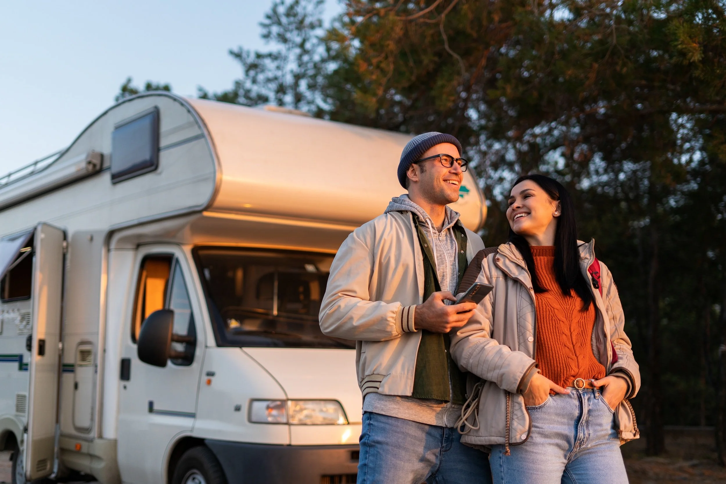 A man and woman standing outside near a white camper van, smiling and talking, with trees in the background during sunset.