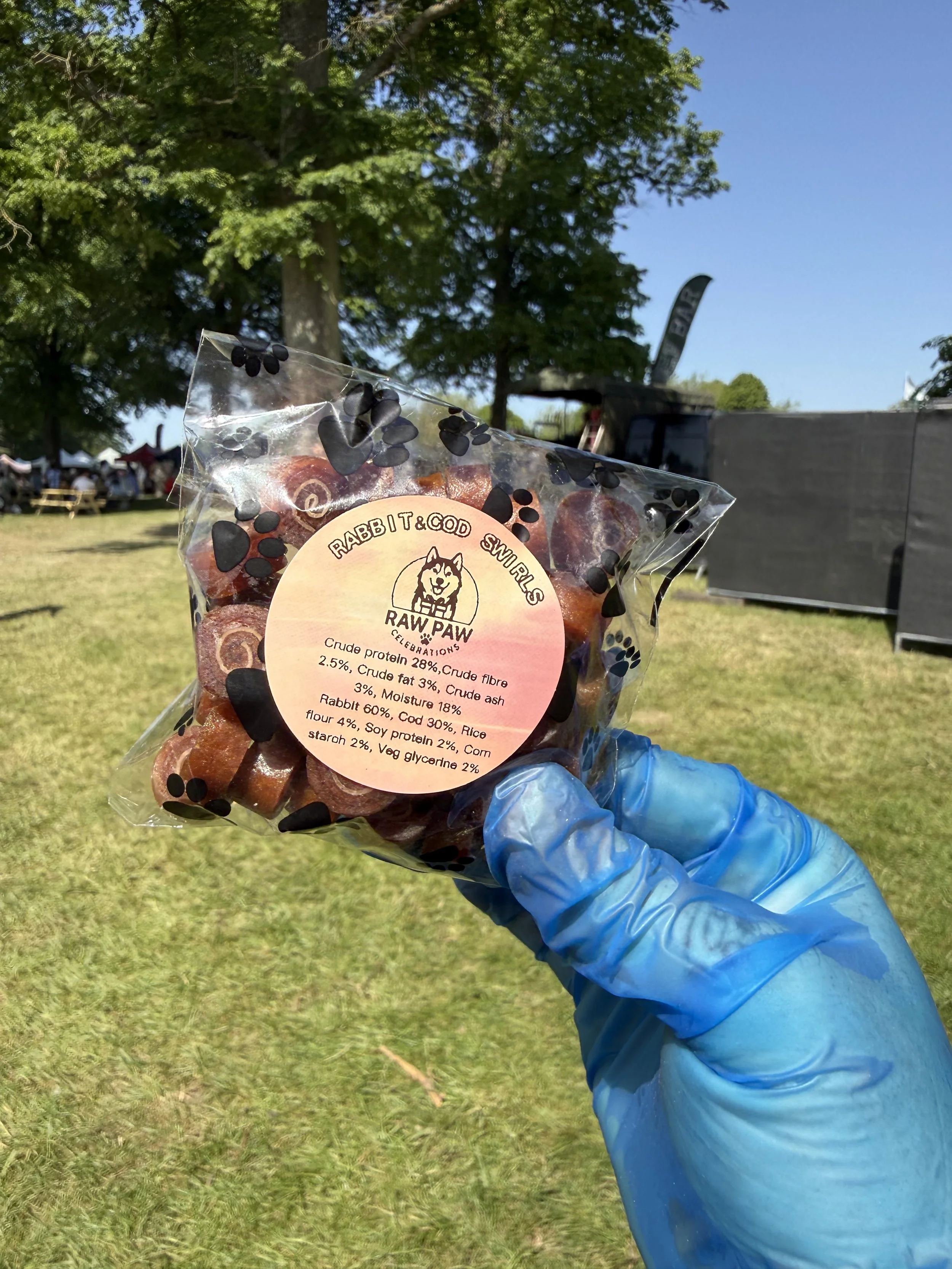 Person in blue gloves holding a clear plastic bag of rabbit and cod snacks at an outdoor event with trees, tents, and people in the background under a blue sky.