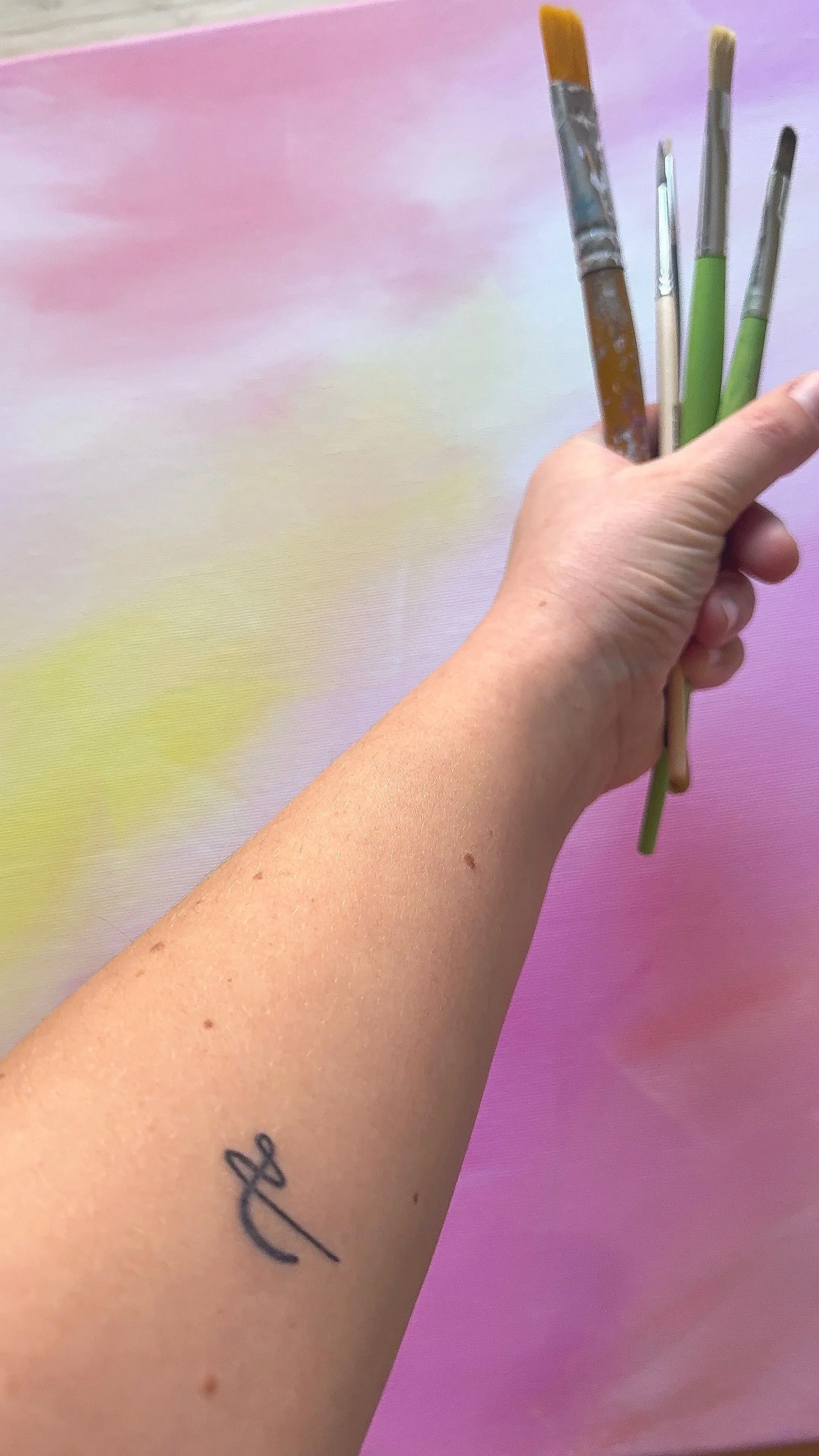 Eine Hand hält mehrere Pinsel vor einem bunten, pastellfarbenen Hintergrund. Auf dem Arm ist ein Tattoo.
