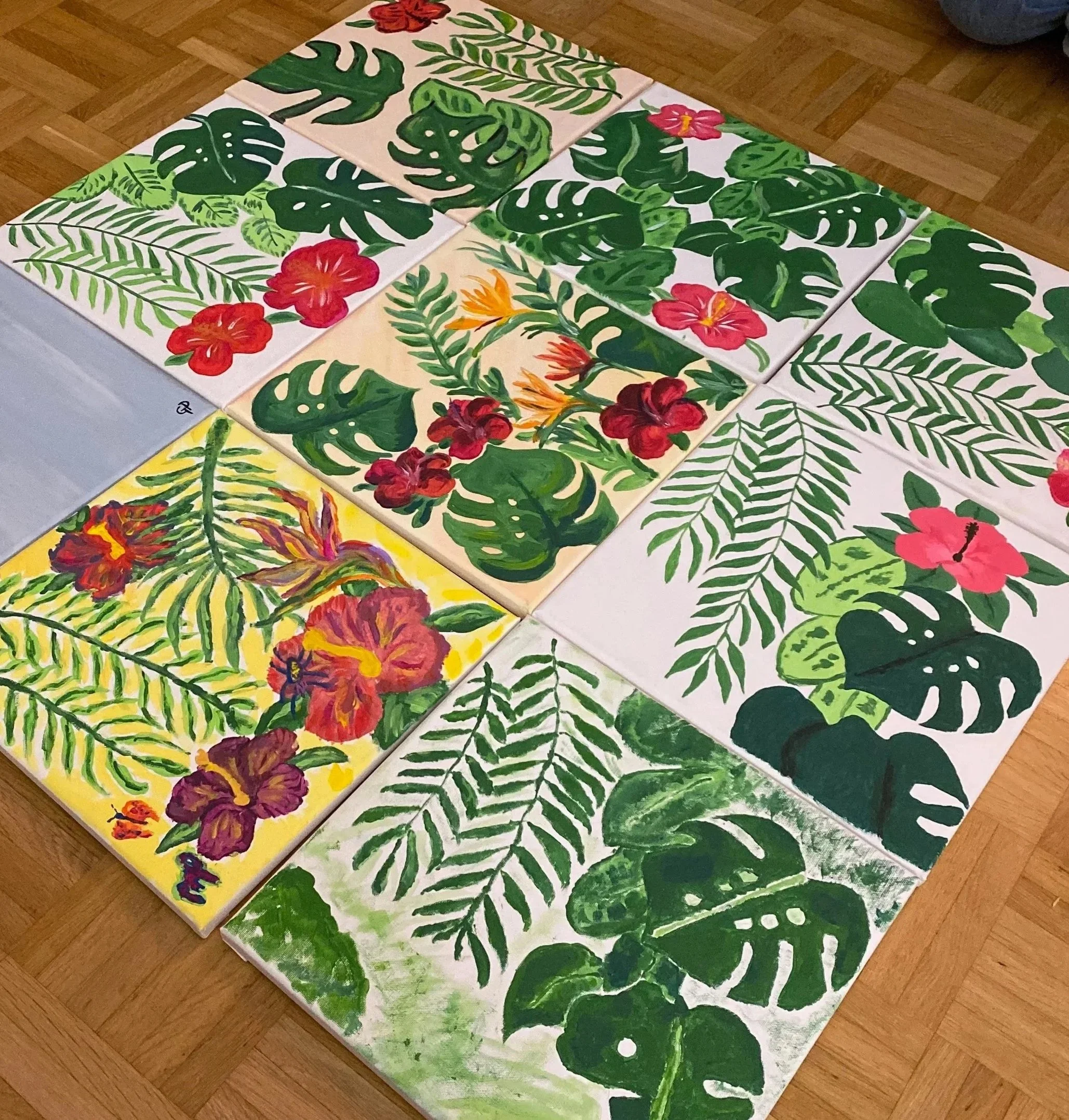 Mehrere bunte Gemälde mit tropischen Pflanzen und Blüten, darunter Monstera- und Farnblätter, auf einem Holzboden ausgelegt.