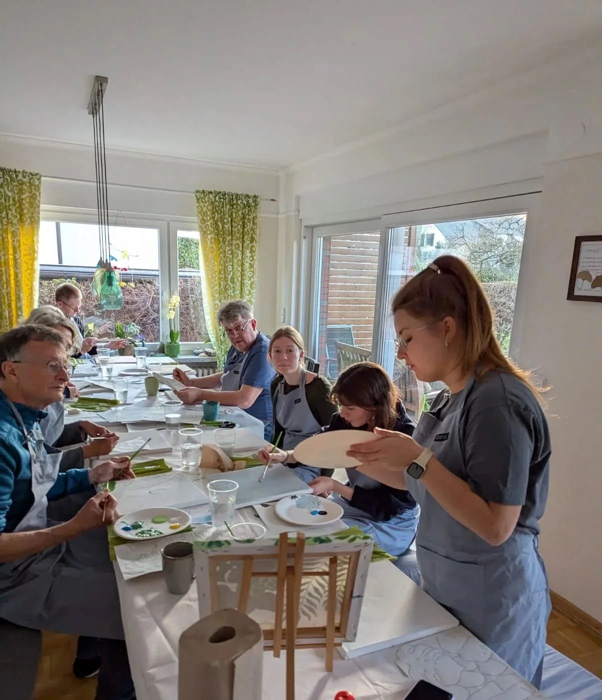 Menschen beim Basteln in einem hell erleuchteten Raum mit großen Fenstern und gelben Vorhängen.