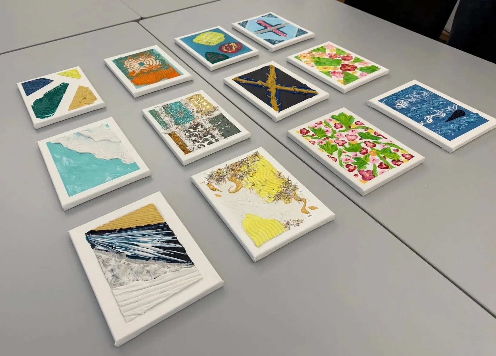 Mehrere kleine künstlerische Gemälde auf weißen Leinwänden, die auf einem grauen Tisch ausgestellt sind. Die Bilder sind vielfältig in Farben und Mustern, darunter Blumenmuster, Wellen, abstrakte Formen und Landschaften.