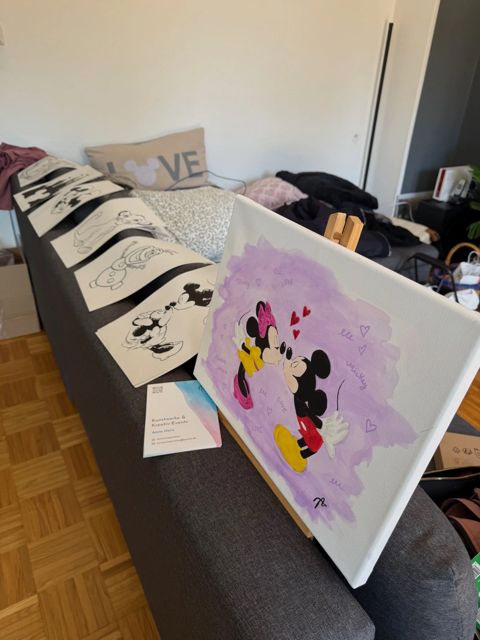 Auf einem grauen Sofa sind mehrere Kunstwerke und Zeichnungen, darunter eine bunte Zeichnung von Mickey und Minnie Mouse, sowie schwarze und weiße Bleistiftzeichnungen von Mickey Mouse in verschiedenen Posen. Im Hintergrund befinden sich Kissen, eine