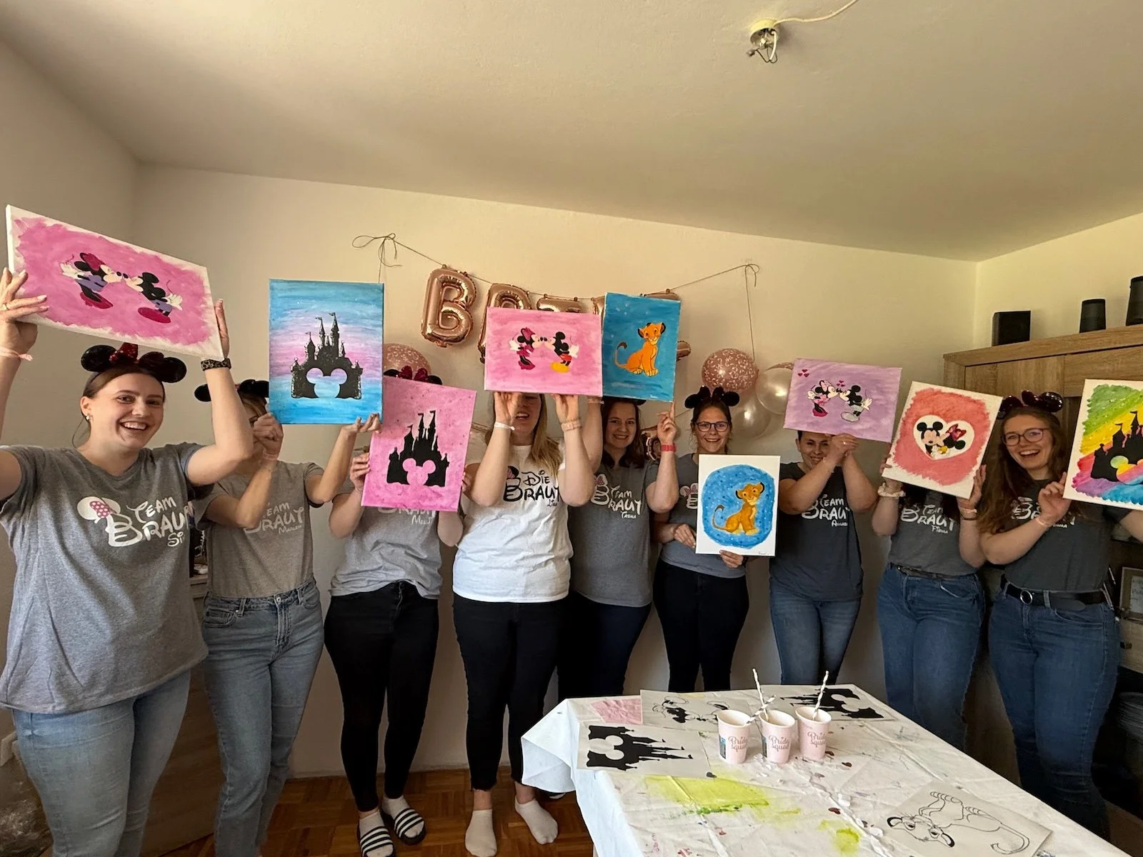 Mehrere Frauen stehen in einem Raum, halten Disney-Themenmalereien mit Mickey Mouse, Dschungelbuch und Löwe hoch, und feiern eine Party mit Ballons und Dekoration.