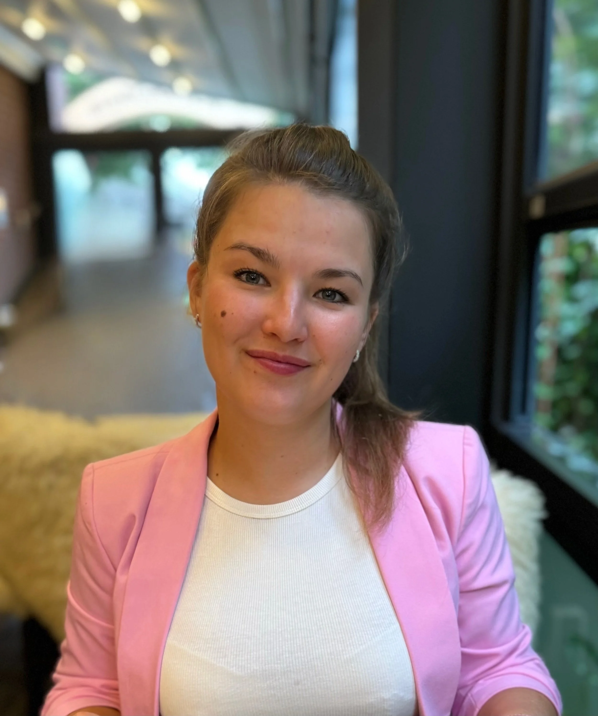 Junge Frau mit braunen Haaren, trägt pinkes Jackett und weißes T-Shirt, sitzt in einem Raum mit großen Fenstern im Hintergrund.