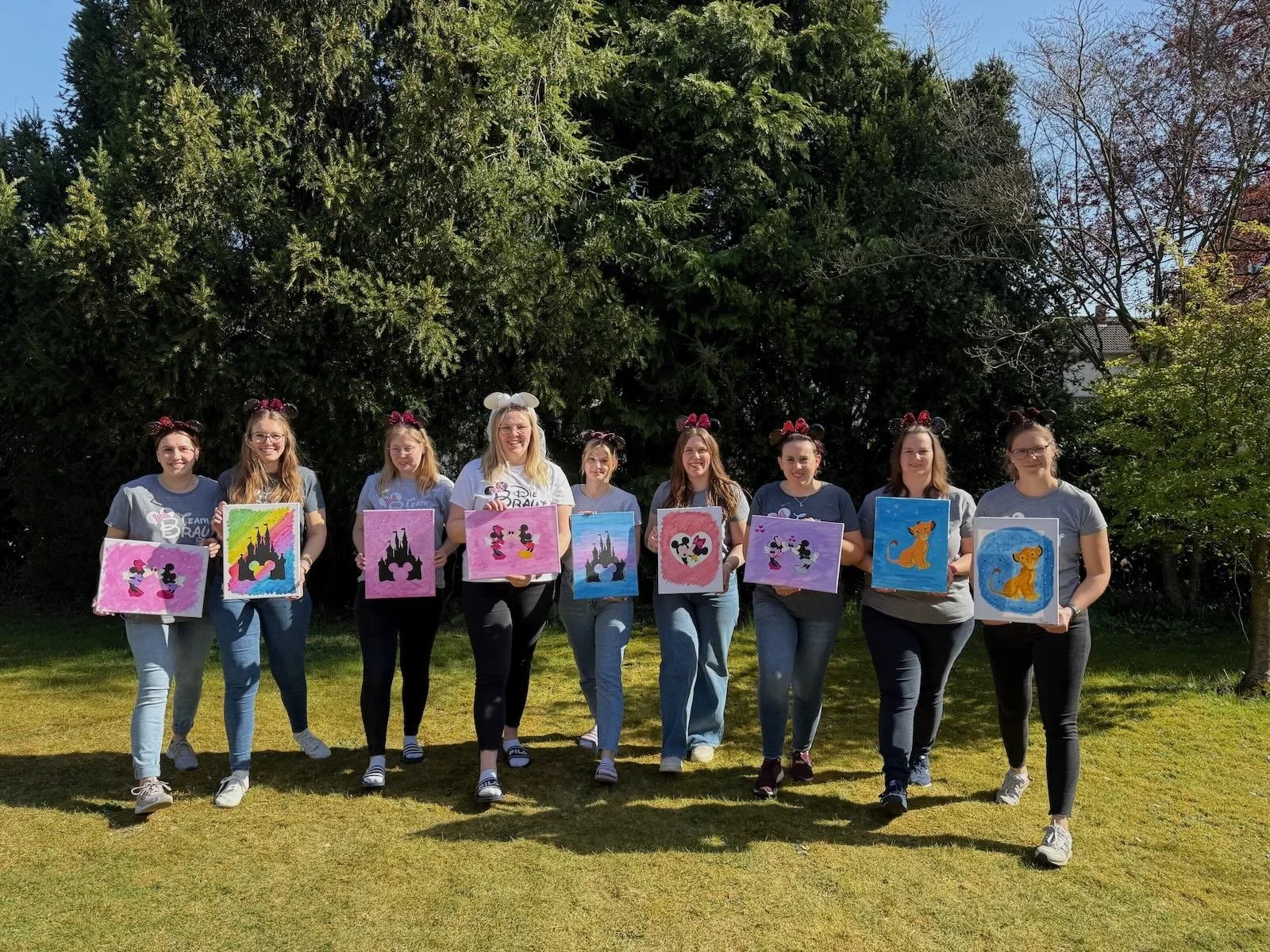 Eine Gruppe von zehn Frauen steht im Garten, alle tragen Minnie Mouse Ohren und halten Gemälde mit Disney-Motiven. Es ist sonnig, und im Hintergrund sind Bäume und ein blauer Himmel sichtbar.