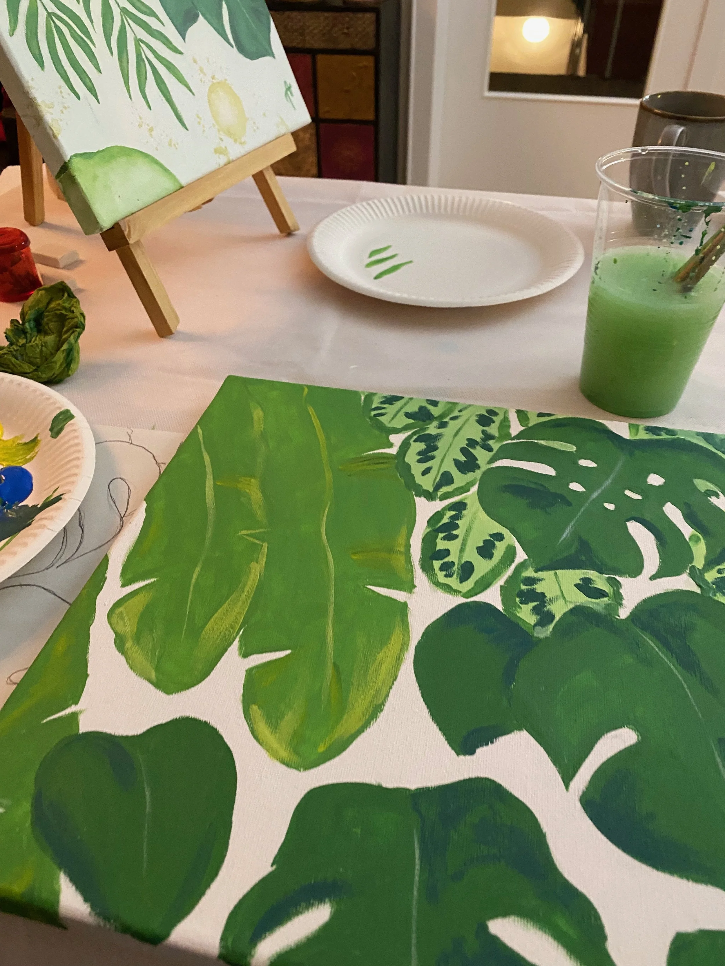 Ein Tisch mit einer weißen Tischdecke, auf der sich eine mit grünen Blättern bemalte Leinwand befindet. Im Hintergrund sind Papierteller mit grünen Pinselstrichen, ein Glas mit grüner Farbe und ein kleiner Malblock zu sehen. Es scheint eine Malaktivi