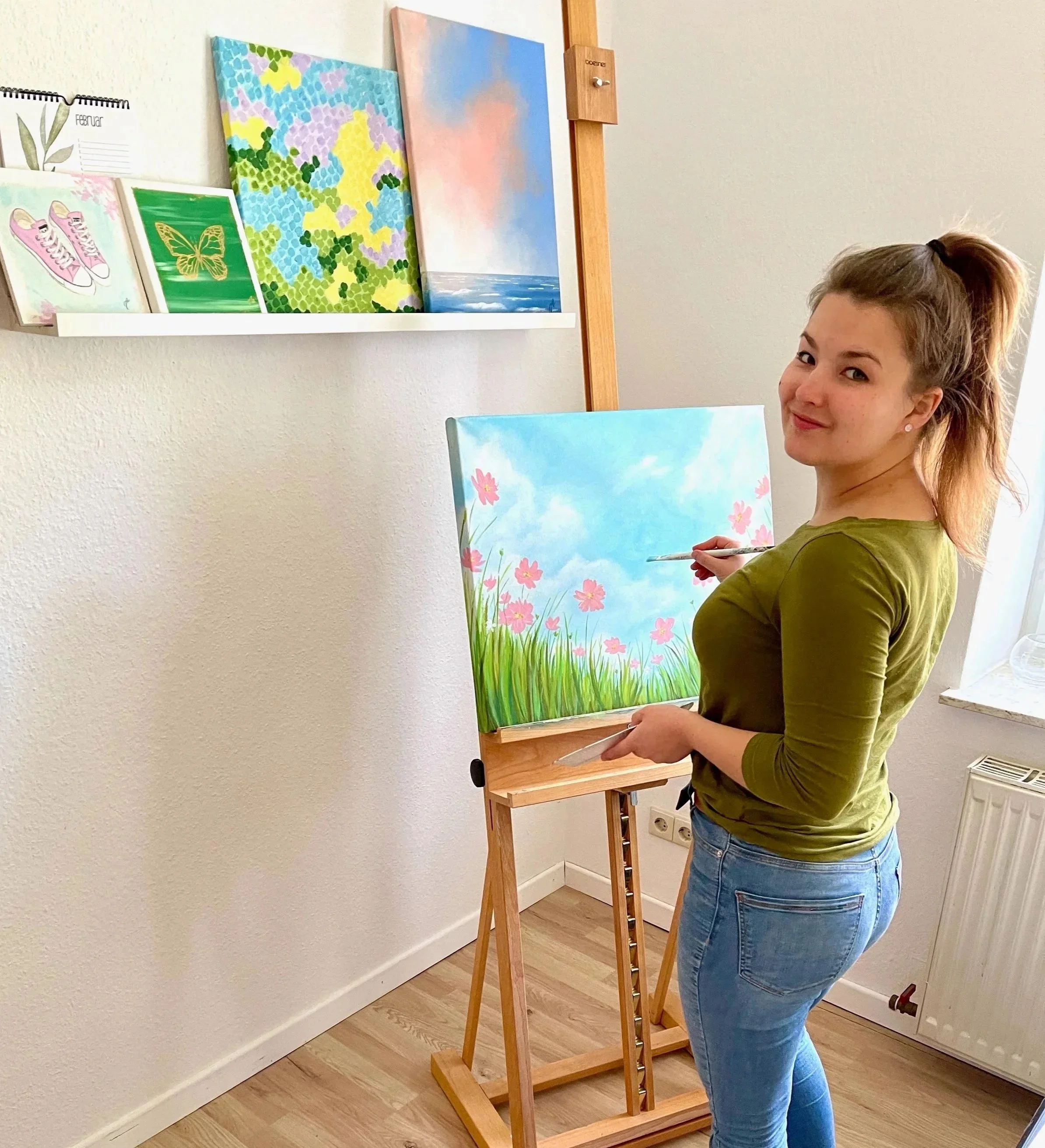 Eine junge Frau, Künstlerin,  beim Malen von einem Kunstwerk vor einer Staffelei. Auf dem Bild ist ein  Himmels mit rosa Blumen zu sehen. In dem Raum sieht man noch weitere künstlerischen Bildern gemalt mit Acryl.