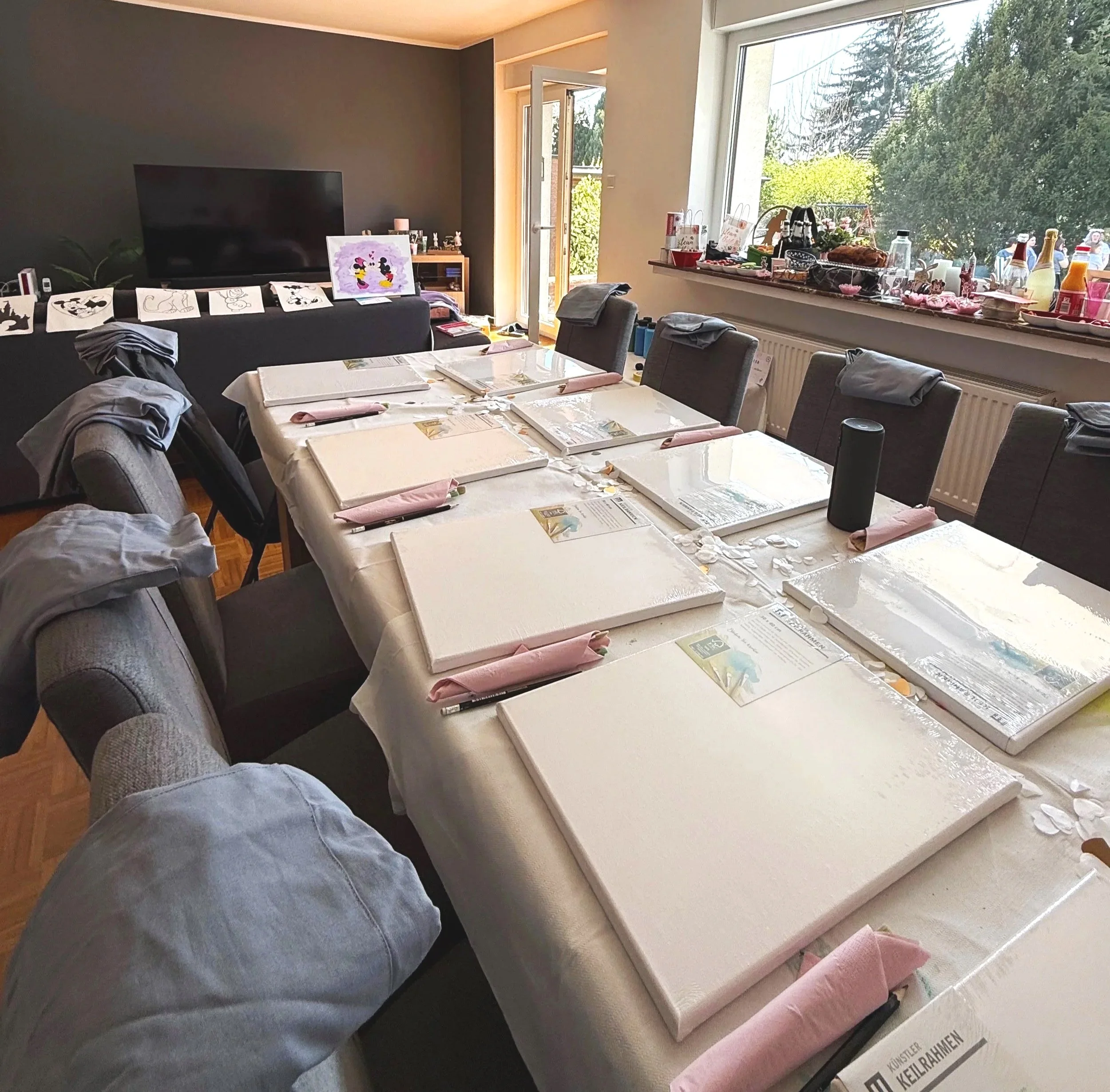 Ein festlich gedeckter Tisch für eine kreative Gruppenaktivität, Malkurs oder Workshop mit Leinwänden, farbigen Servietten, Pinseln und einer hellen Tischdecke  an einem großen Fenster mit Blick auf einen Garten bei Tageslicht.