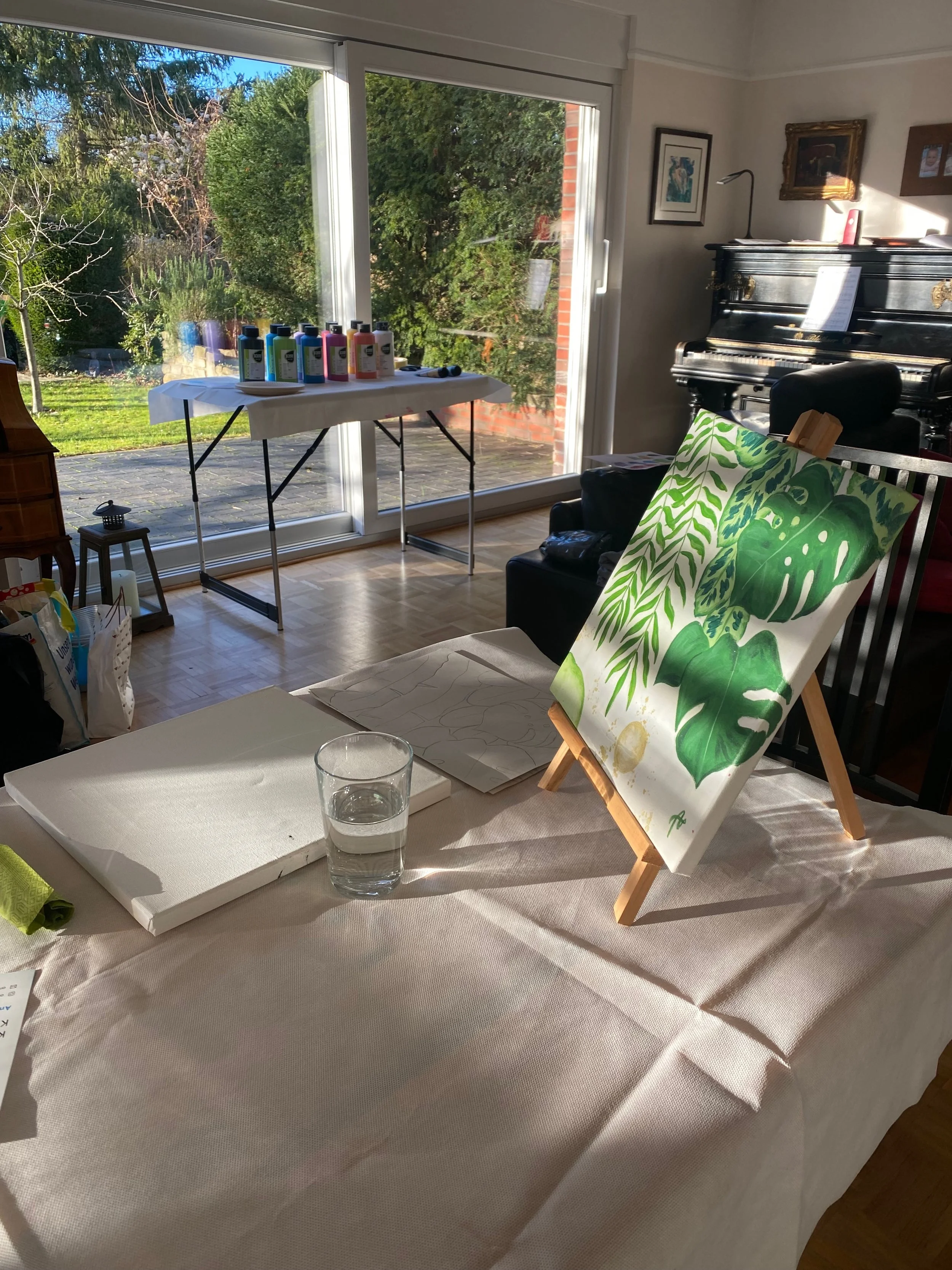 Ein heller Raum mit großen Fenstern, durch die man einen Garten sieht. Auf einem Tisch liegen Malutensilien, auf einem kleinen Ständer steht eine Leinwand mit grünem Pflanzenmuster. Im Hintergrund steht ein schwarzer Klavier. 