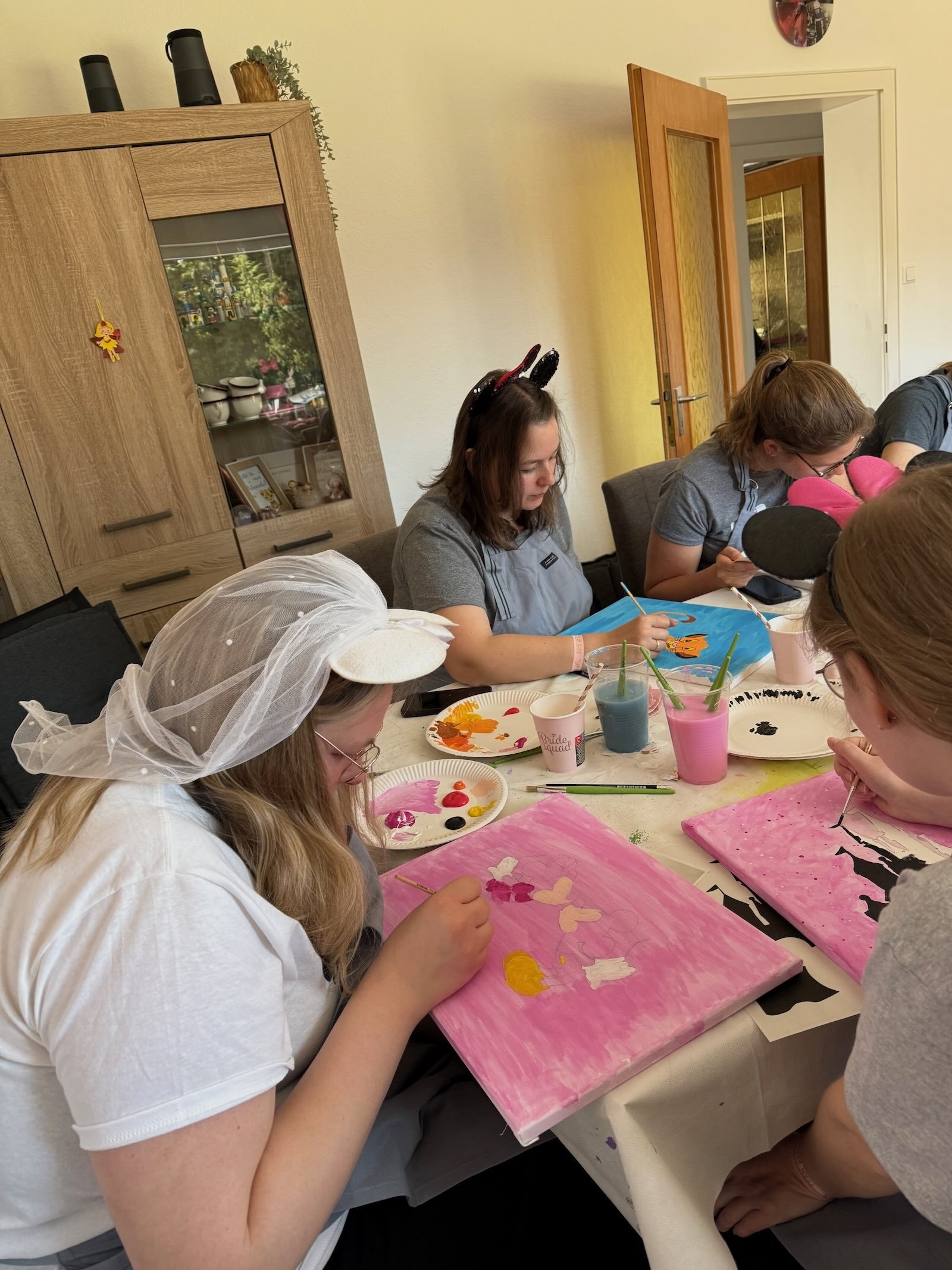 Menschen beim Malen von Kunstwerken auf Keilrahmen, einige tragen dekorative Kopfbedeckungen in einem Raum mit Tisch und Dekoration. Es werden Disneyfiguren gemalt.