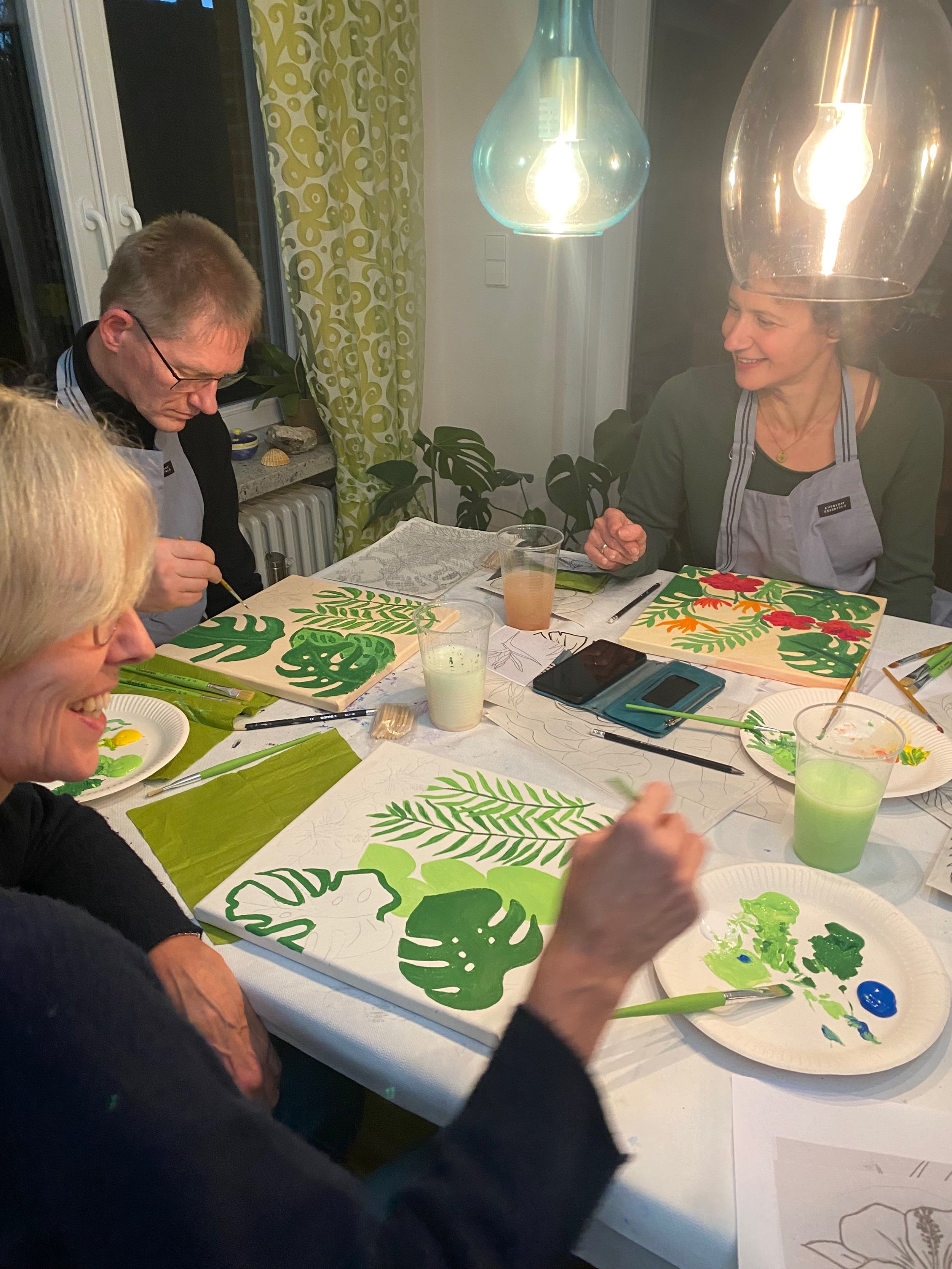 Menschen beim gemeinsamen Malen von Blattmustern mit grüner Farbe in einem Raum bei Kerzenlicht.