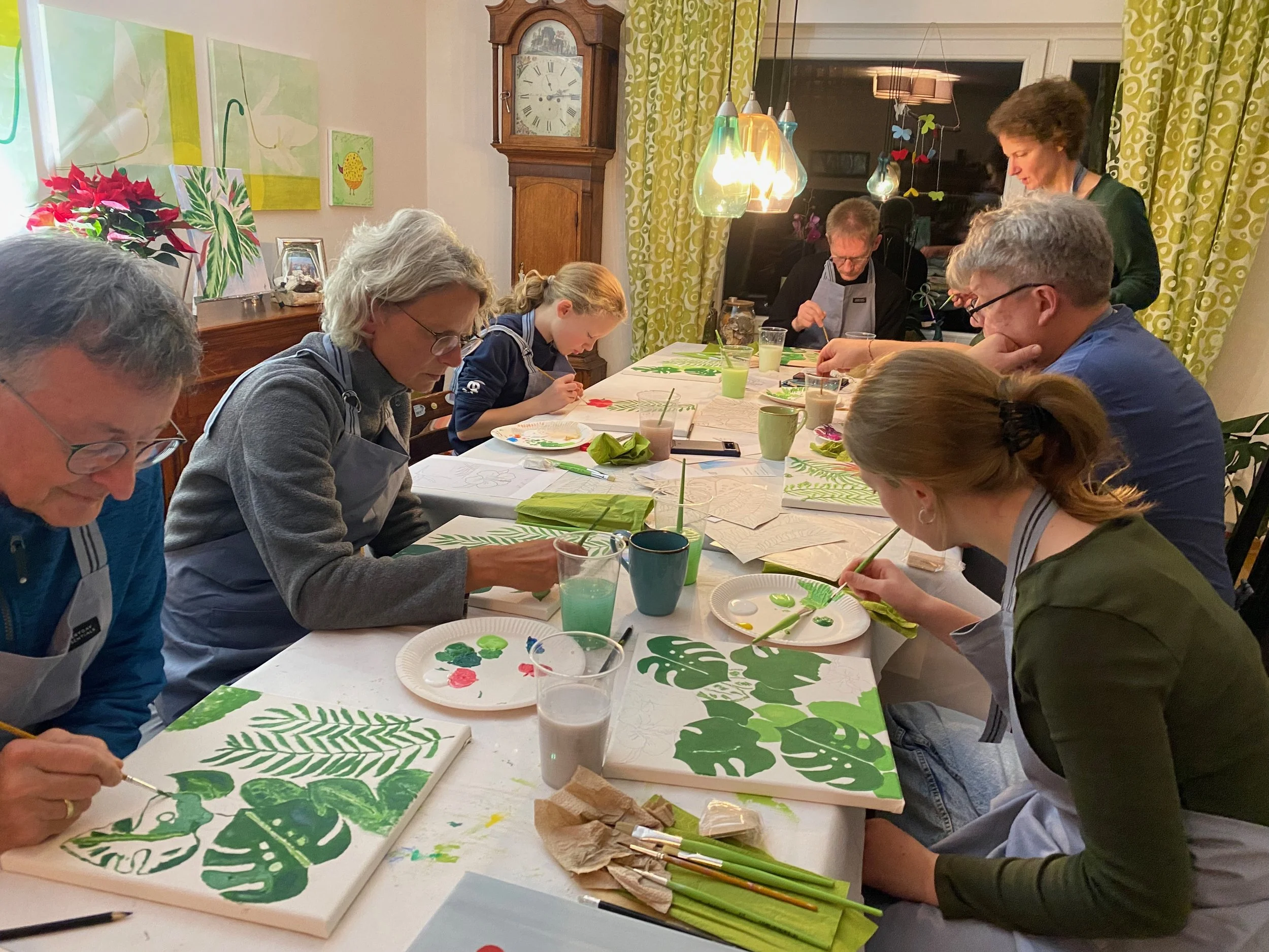 Menschen beim gemeinsamen Malen mit grünen Blattformen auf Leinwänden in einem Raum mit Holzmöbeln und grünen Vorhängen.