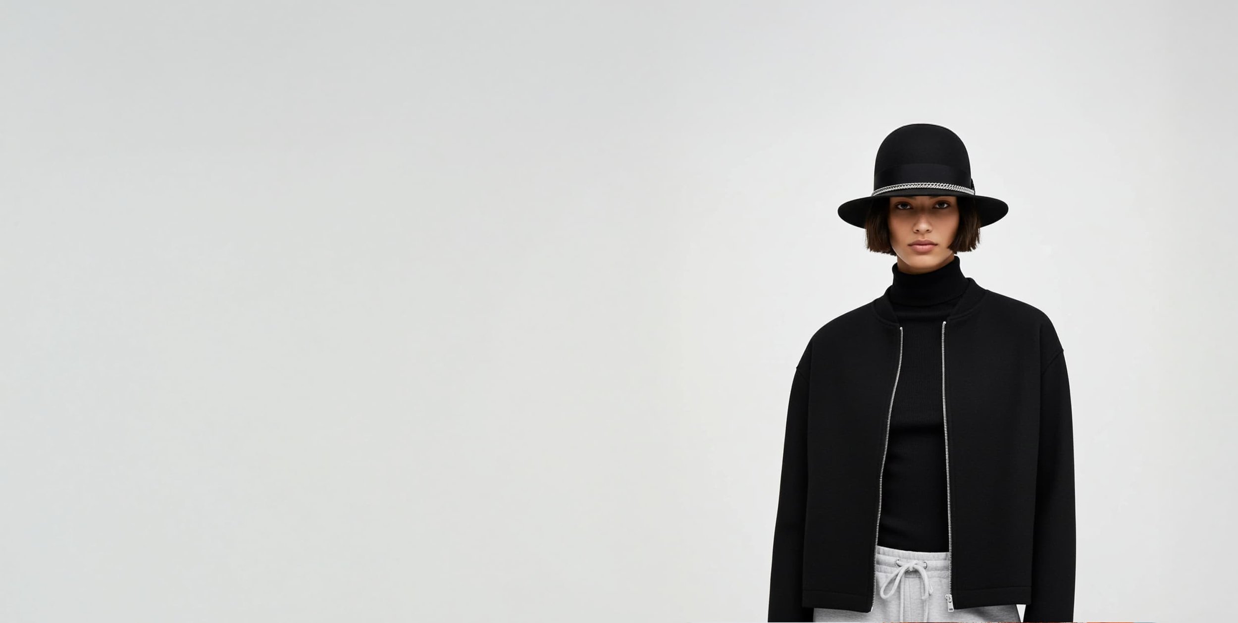 A woman wearing a black hat, black jacket, black turtleneck, and gray sweatpants against a plain white background.