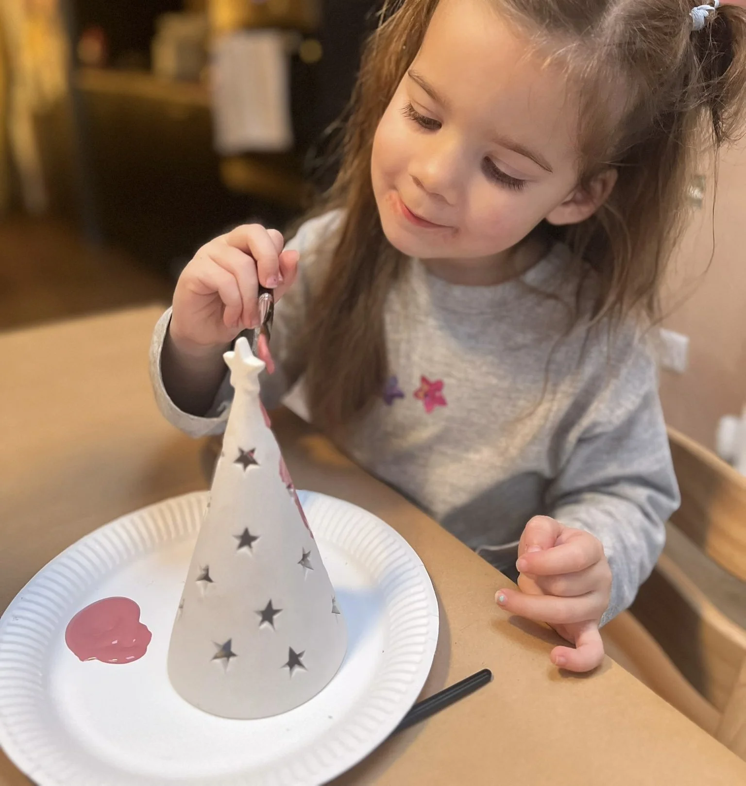 A young girl with long hair, wearing a gray sweater, is decorating a white cone-shaped Christmas tree lantern. She is using a small paintbrush and has a subtle smile. The lantern is placed on a white paper plate with pink paint on it.