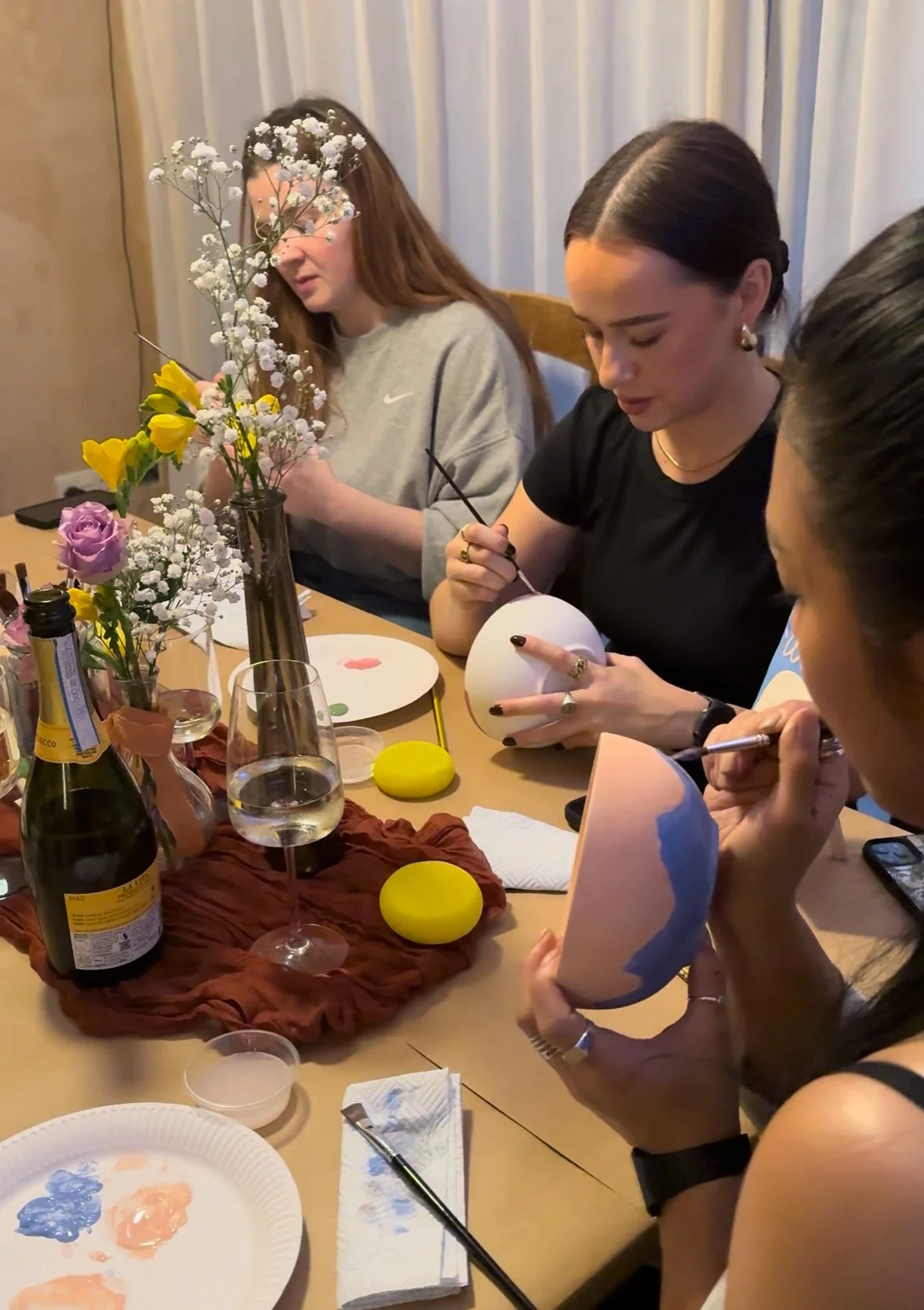 Four women sit at a table decorating pots with paint and brushes, with flowers and art supplies on the table.