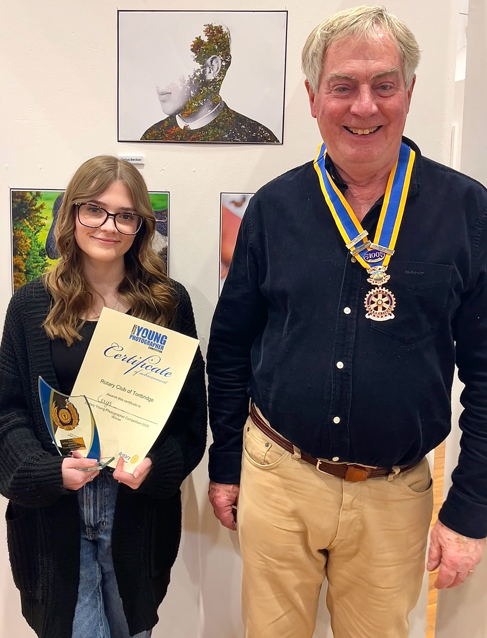 A young woman holding a certificate and a trophy standing next to an older man wearing medals around his neck, in front of artworks on a wall.