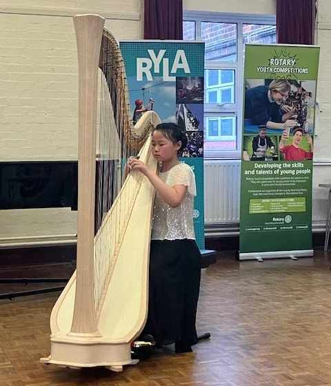 A young girl playing a large wooden harp indoors near two vertical banners, one with the acronym 'RYLA' and the other promoting Rotary Youth Competitions and developing young people's skills.