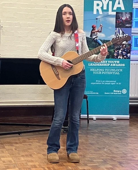 A young woman with long dark hair playing an acoustic guitar on stage during an event, with a blue RYLA banner in the background.