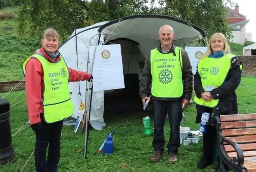 Three people wearing Rotary Club vests standing outdoors in front of a tent, with trees in the background, during daytime.